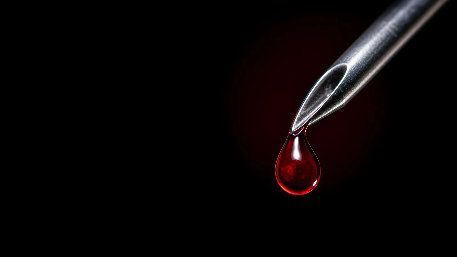 A single drop of crimson blood on the polished bevel of a steel needle, catching the light against black.